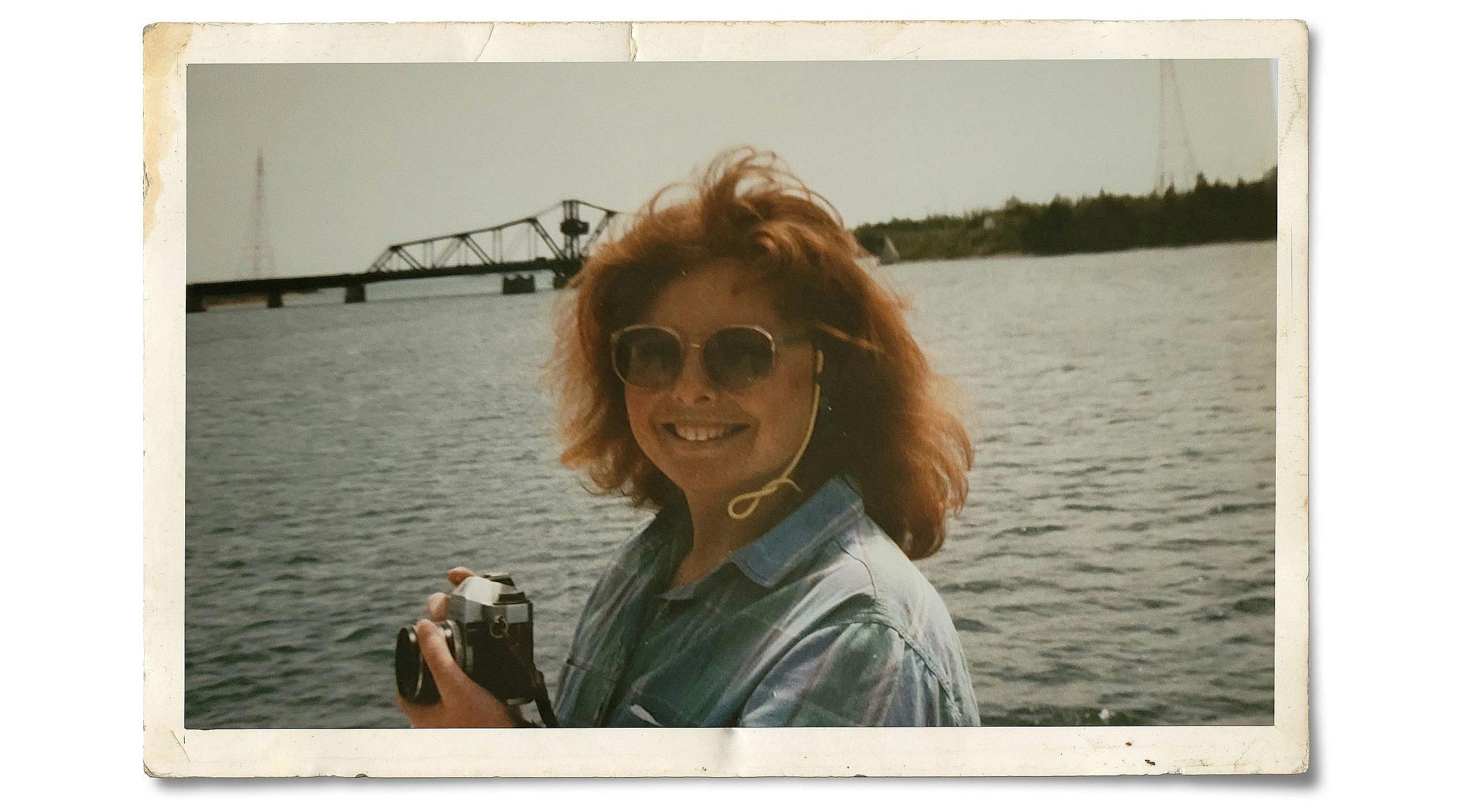 Woman with short orange hair and wearing a washed out blue shirt and black sunglasses holding a camera, smiling, against the backdrop of a large body of water.