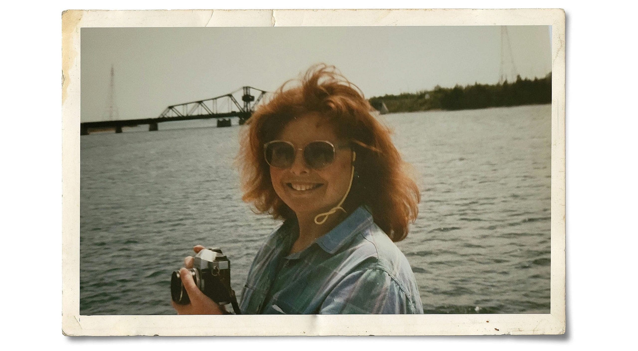 Woman with short orange hair and wearing a washed out blue shirt and black sunglasses holding a camera, smiling, against the backdrop of a large body of water.