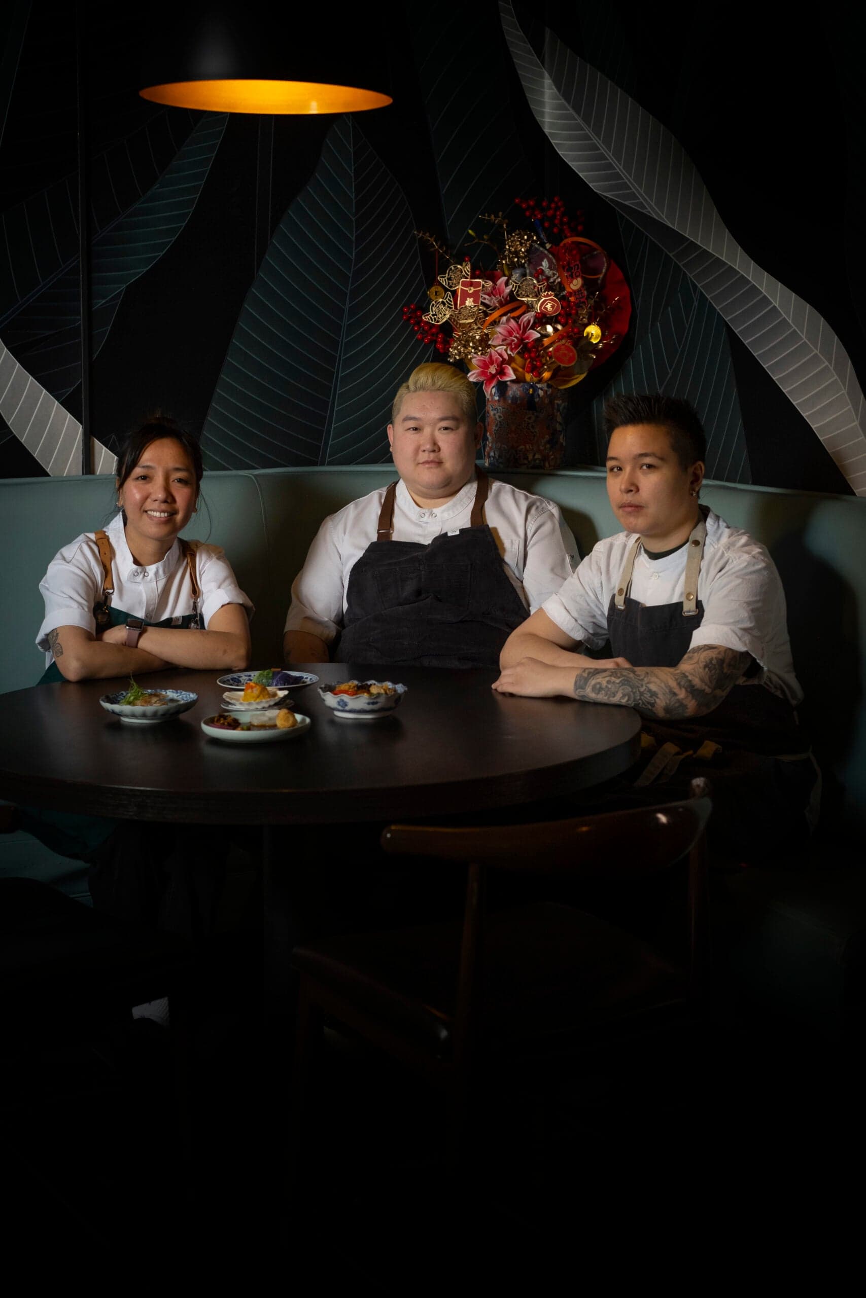 Three chefs in white t-shirts and black aprons, sitting at a table set with Chinese food