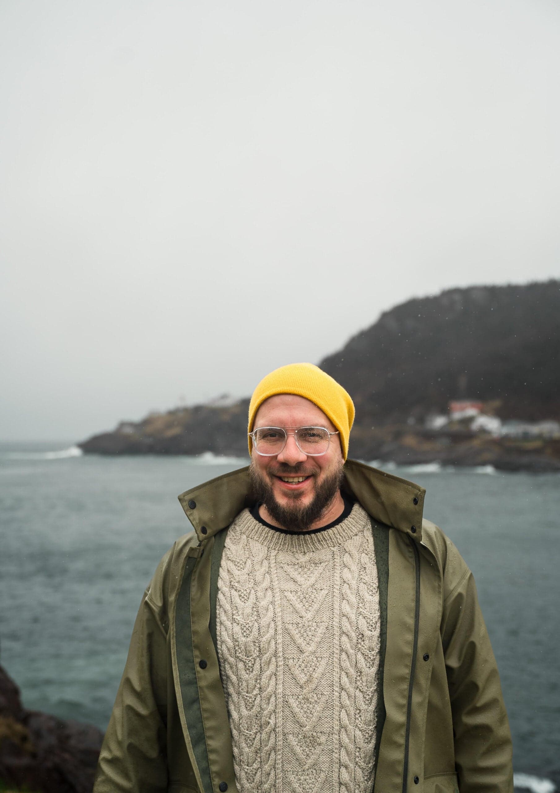 A man in a fisherman's sweater and white beanie standing by the coast