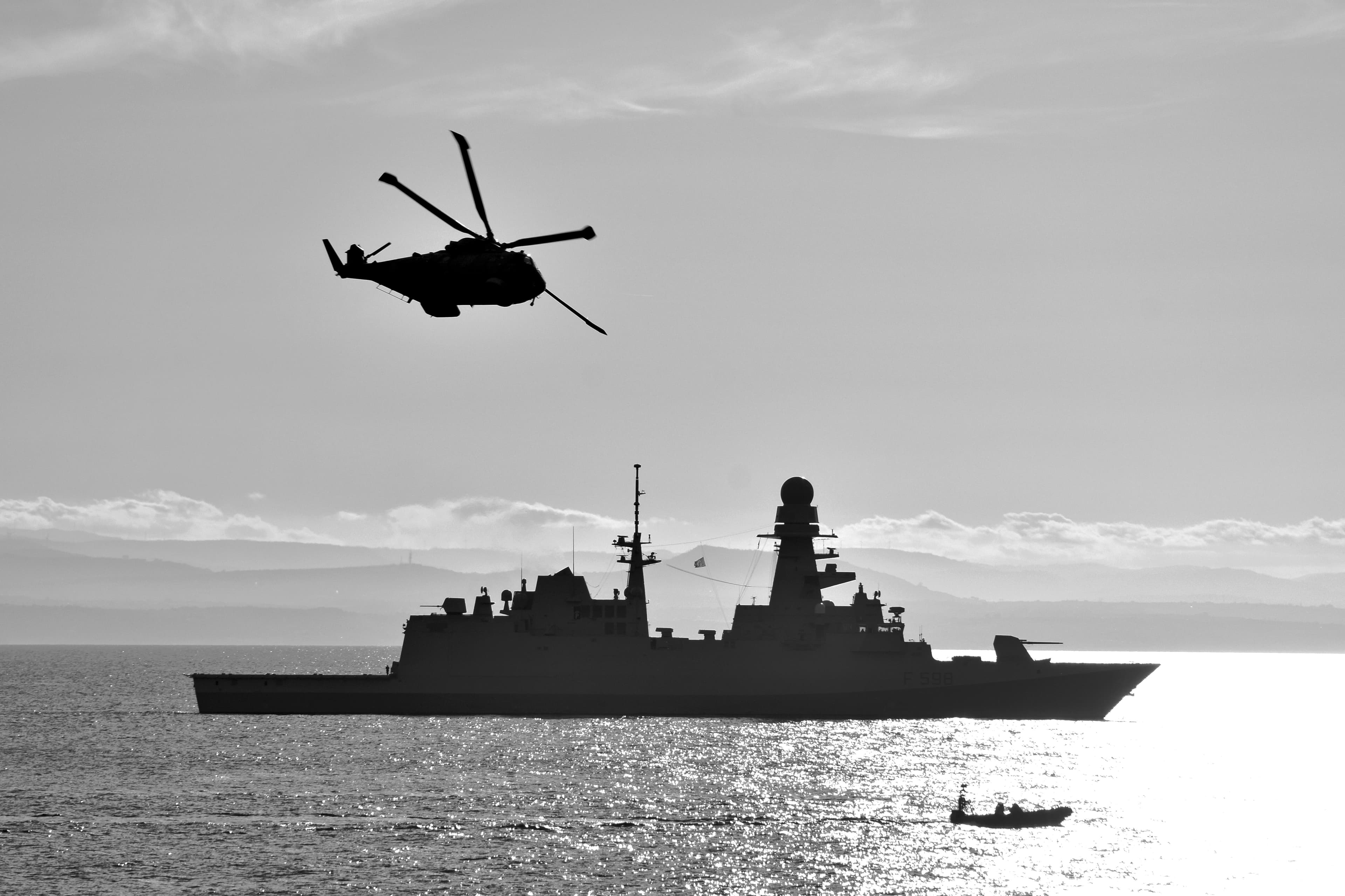 A military helicopter flying above a naval ship, photographed in black and white