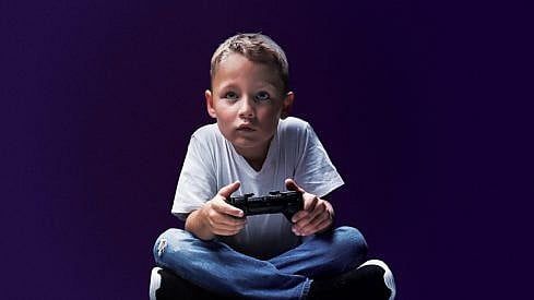 A young boy in a white t-shirt and blue jeans sits cross-legged and stares intently ahead, holding a gaming console in his hand