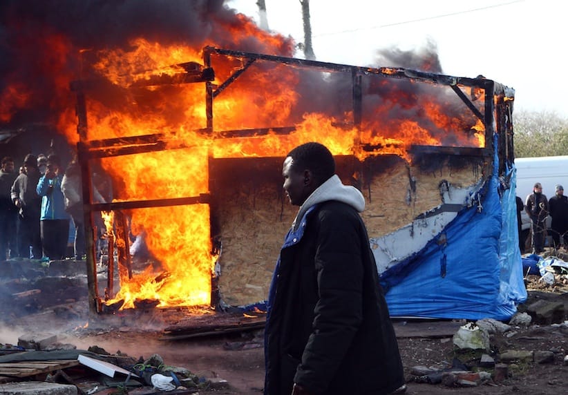 A migrant walks past a burning dwelling in the makeshift migrants camp near Calais, France in March, 2016. A destruction crew on Wednesday, March 16 tore down the last of more than 1,000 tents and huts in a large swath of a makeshift migrant camp in the French port city of Calais, a job marked by fiery protests, an ongoing hunger strike and several arrests. The prefecture, or state authority, which ordered the dismantling of the southern sector of the camp - a veritable village on 7.5 hectares (18.5 acres) of land - said workers guarded by police finished pulling down the flimsy dwellings at 2 p.m. Wednesday, less than three weeks after the job began.  (AP Photo/Michel Spingler, file)