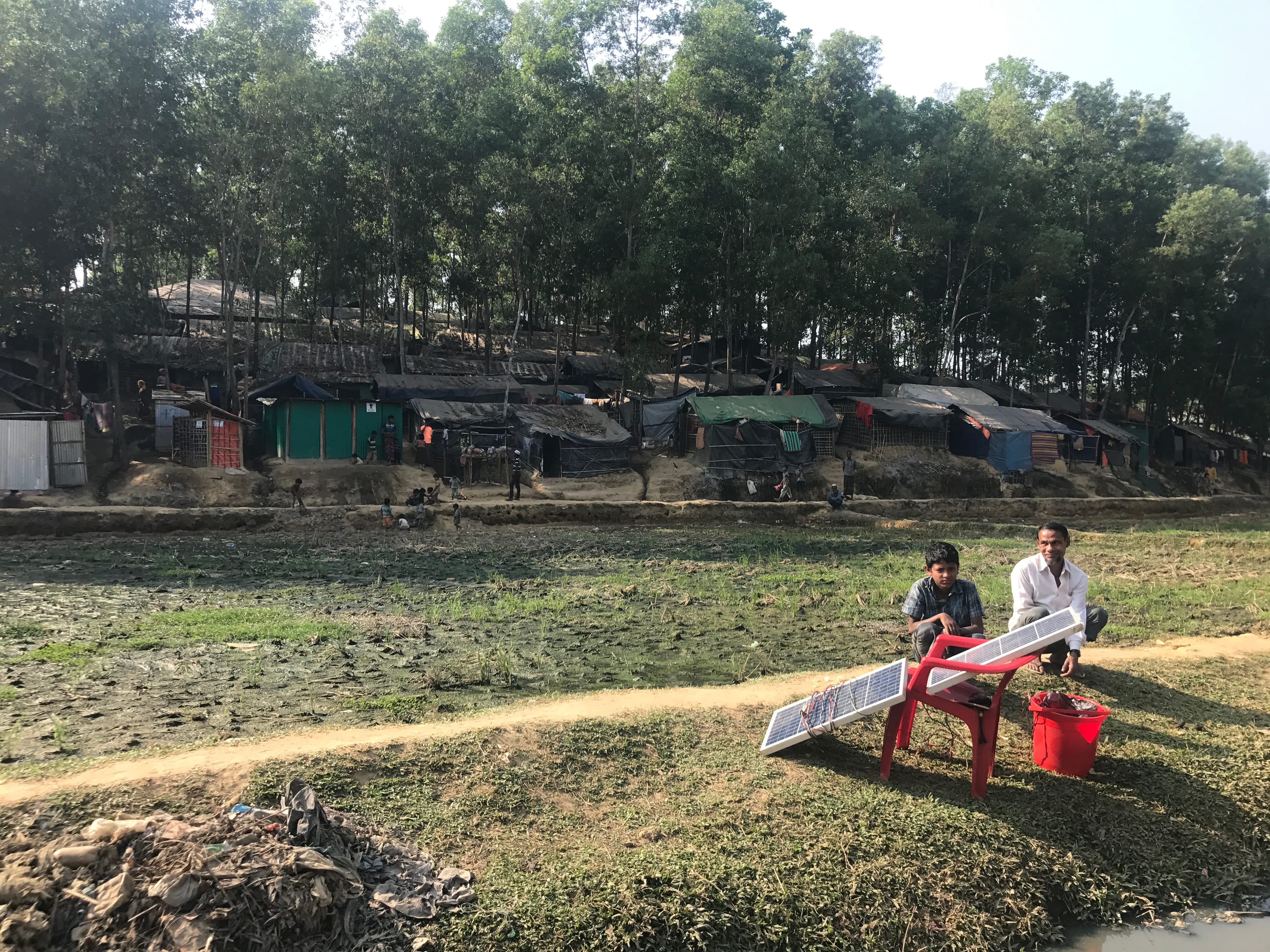 Ten-year-old Ajijur Rahman and his father, Mohammad Siddiq, sit between paddy fields and an irrigation canal by Bangladesh’s Kutupalong Rohingya refugee camp. (Ashish Malhotra)