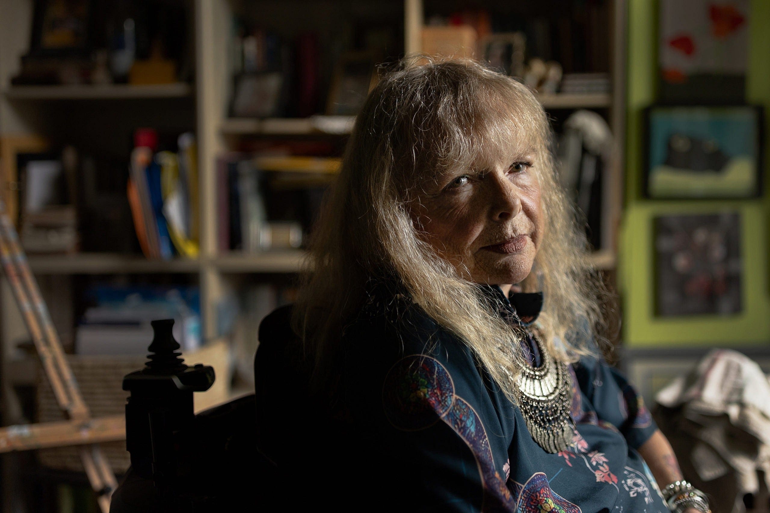 Elderly woman with shoulder length blonde hair looking into the camera. She's wearing a dark blue floral tunic with a double-layered silver necklace