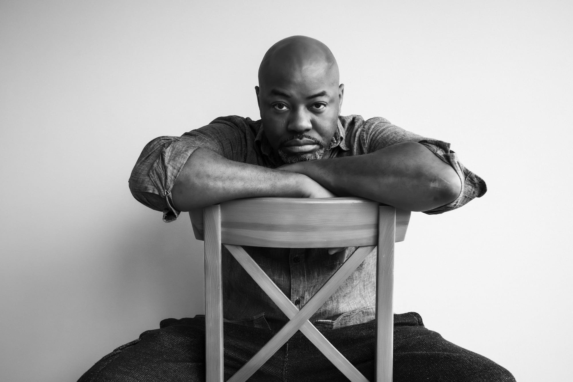 A black and white photo of a man sitting backwards on a wooden chair, his hands resting on his crossed arms which lie on the cahir back.