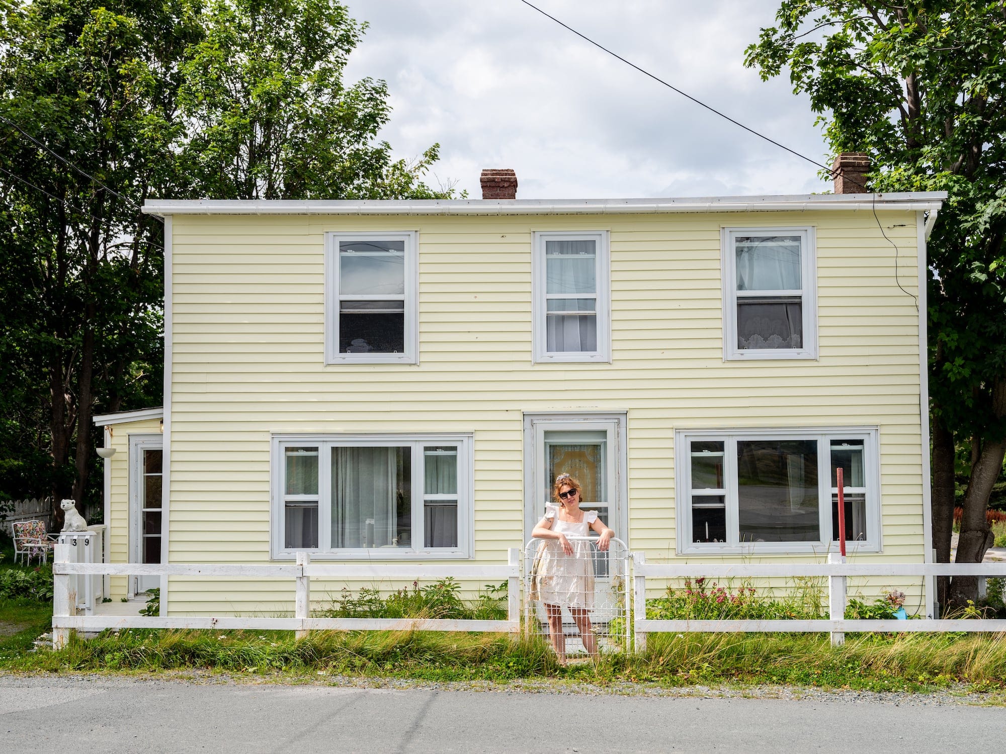 A woman leans against the gate of a white fence. She stands in front of a pale yellow house.