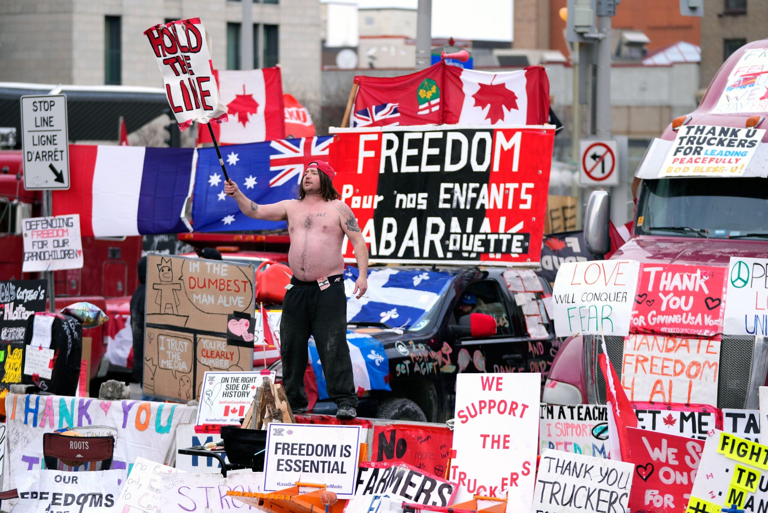 A man standing shirtless holding a picket sign, surrounded by other signs from the freedom truck convoy.