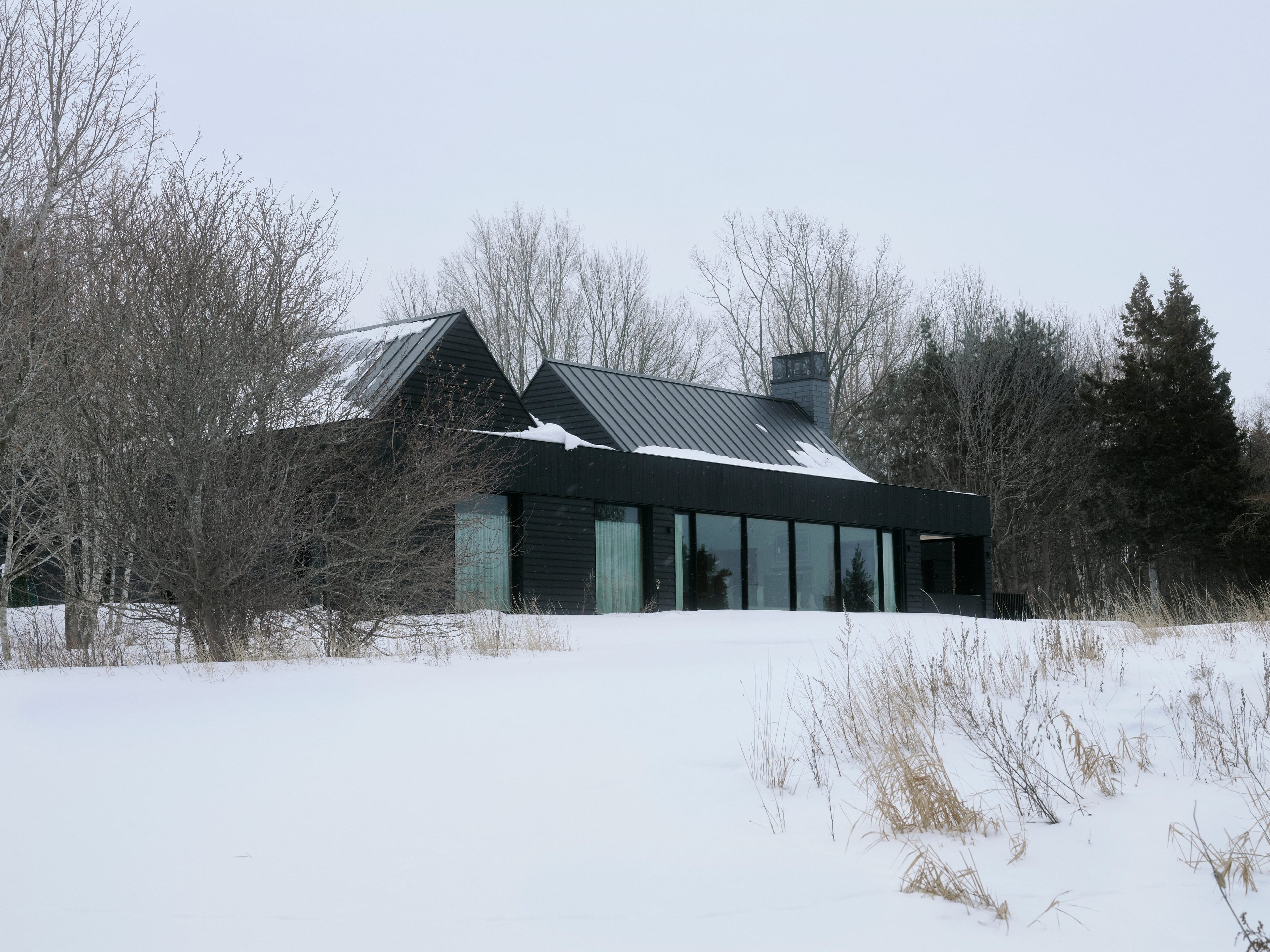 A single-storey black house with large windows and a pitched roof. It is winter and there are trees behind it