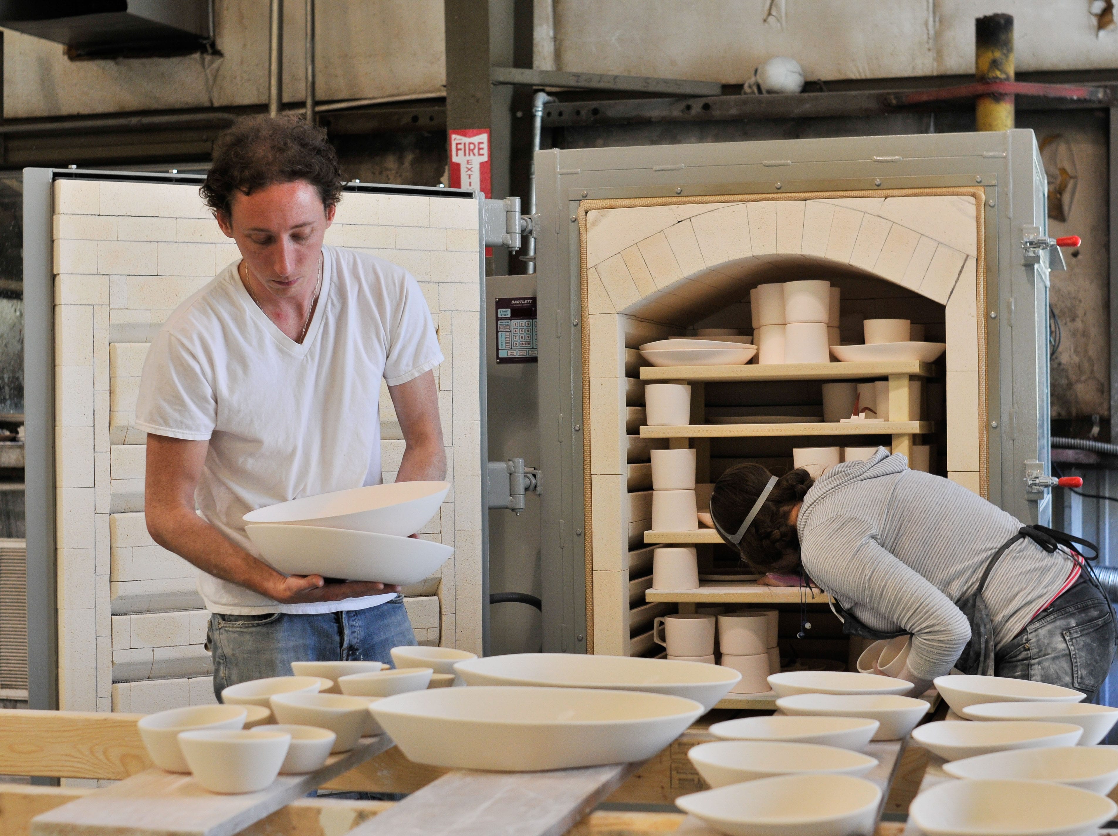 Two ceramic artists loading white bowls, cups, and dishes into large industrial kilns inside a pottery studio.