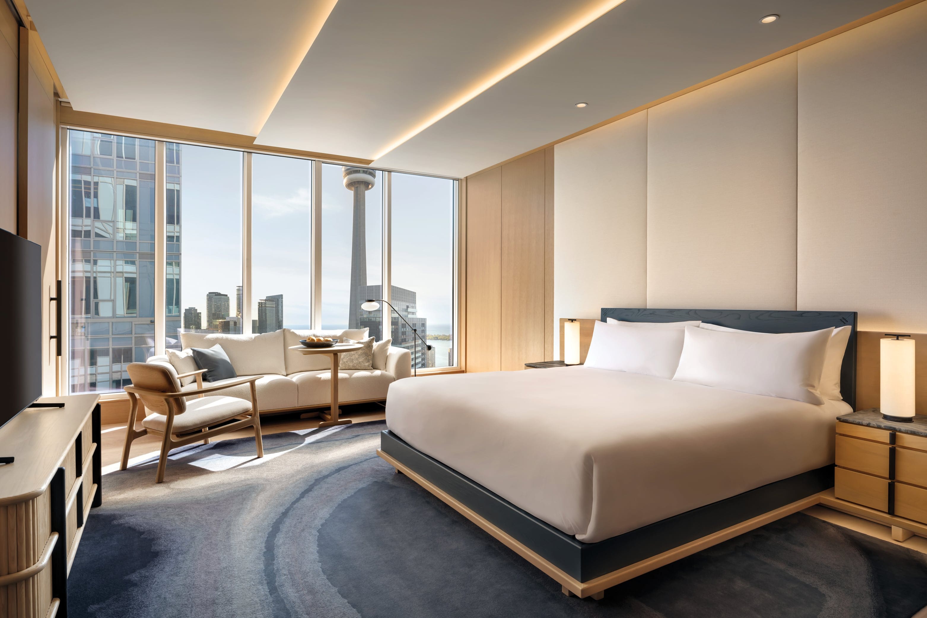 Inside a Nobu Hotel room with warm yellow lighting, a large bed and furnitures are placed on the deep navy carpet. A part of the CN tower and other buildings can be seen from the big window.