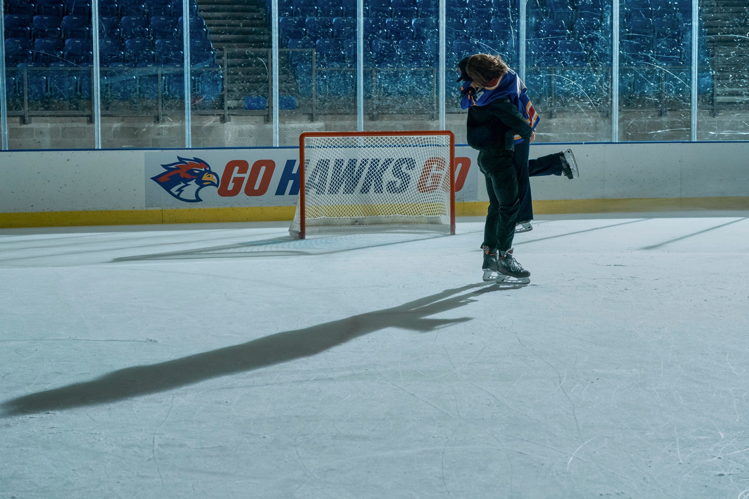 Two people in hockey uniforms and skate shoes are hugging each other on a hockey link.