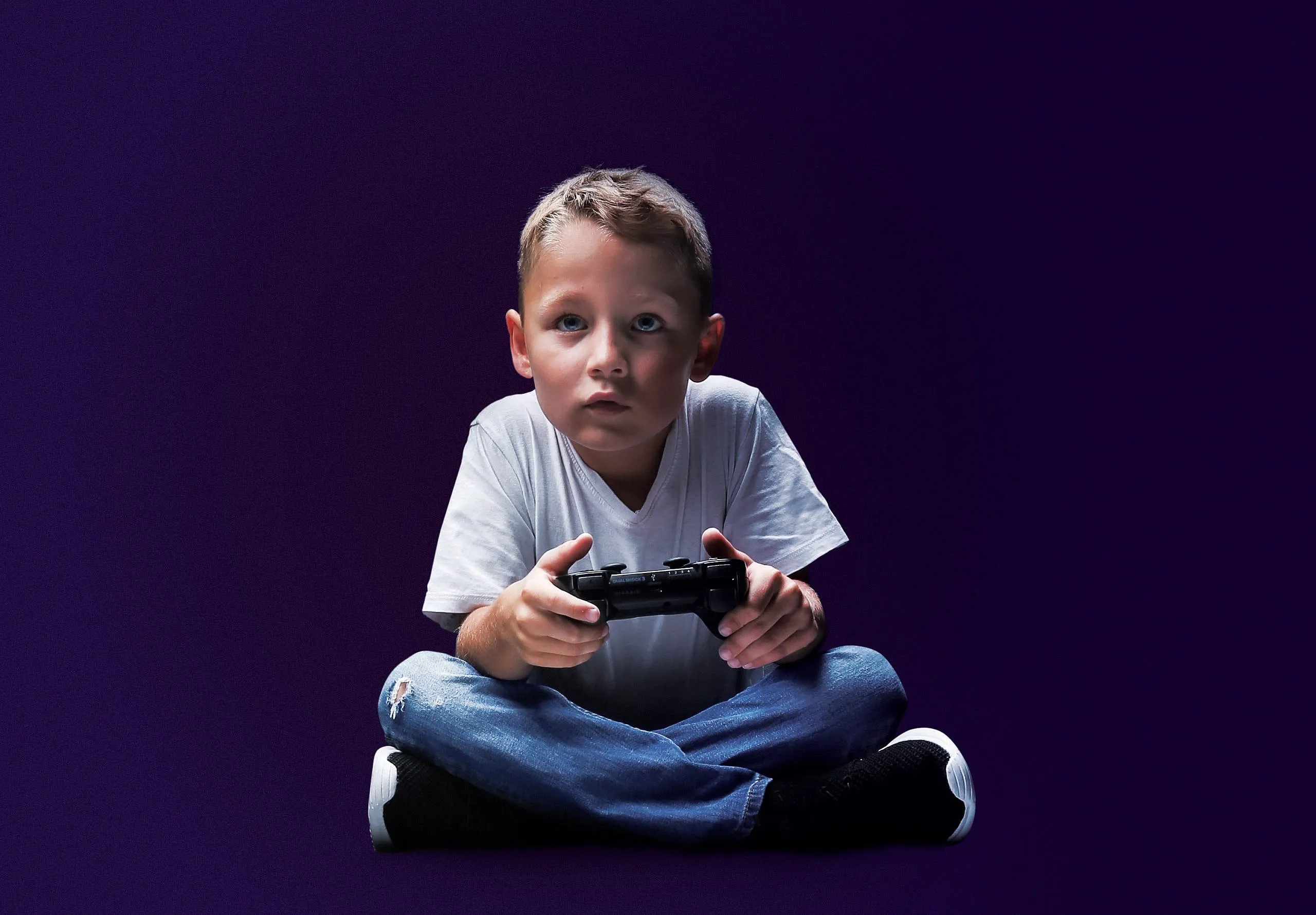 A young boy in a white t-shirt and blue jeans sits cross-legged and stares intently ahead, holding a gaming console in his hand