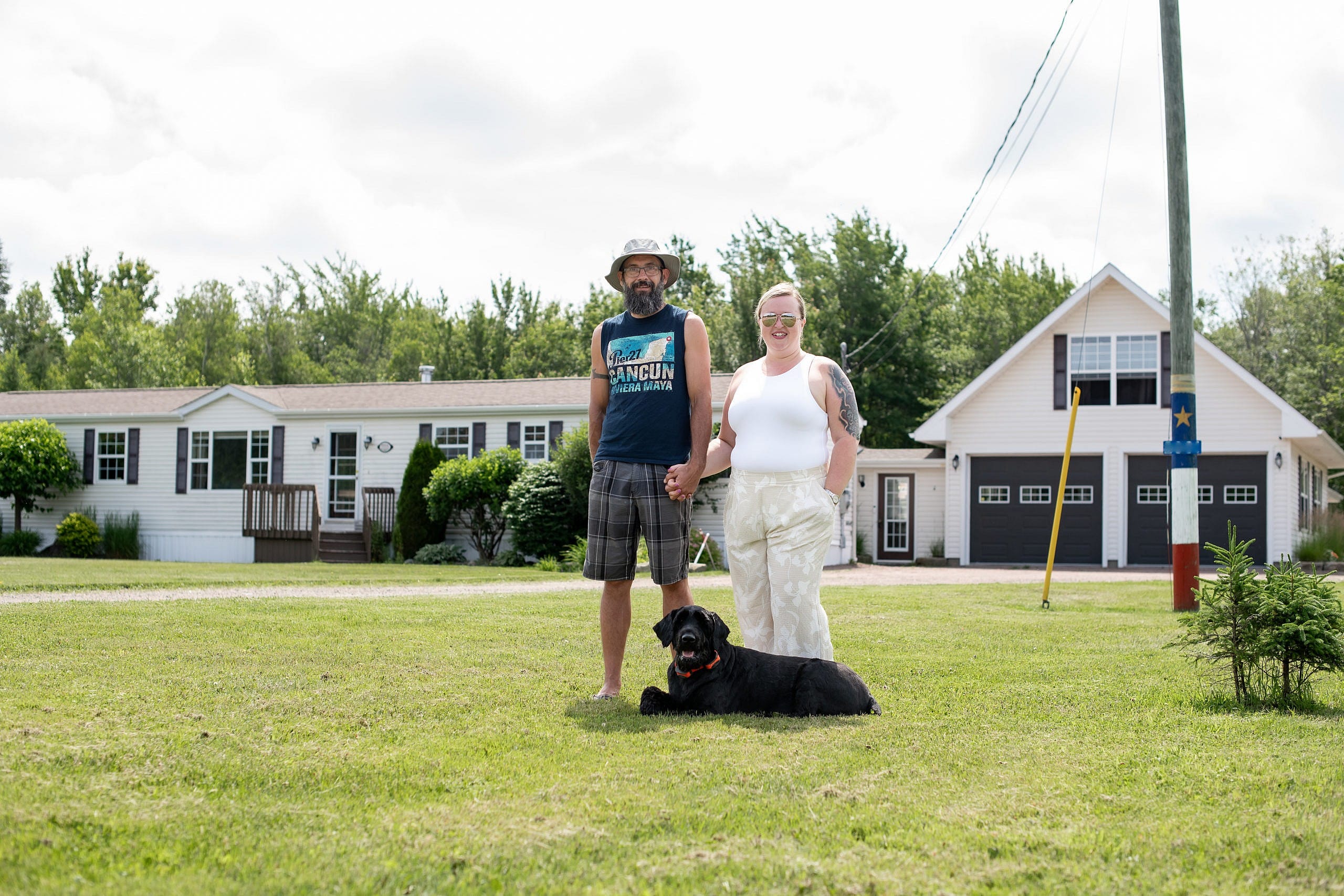 A man and a woman standing and holding hands with a black dog by their feet, standing in front of a white house