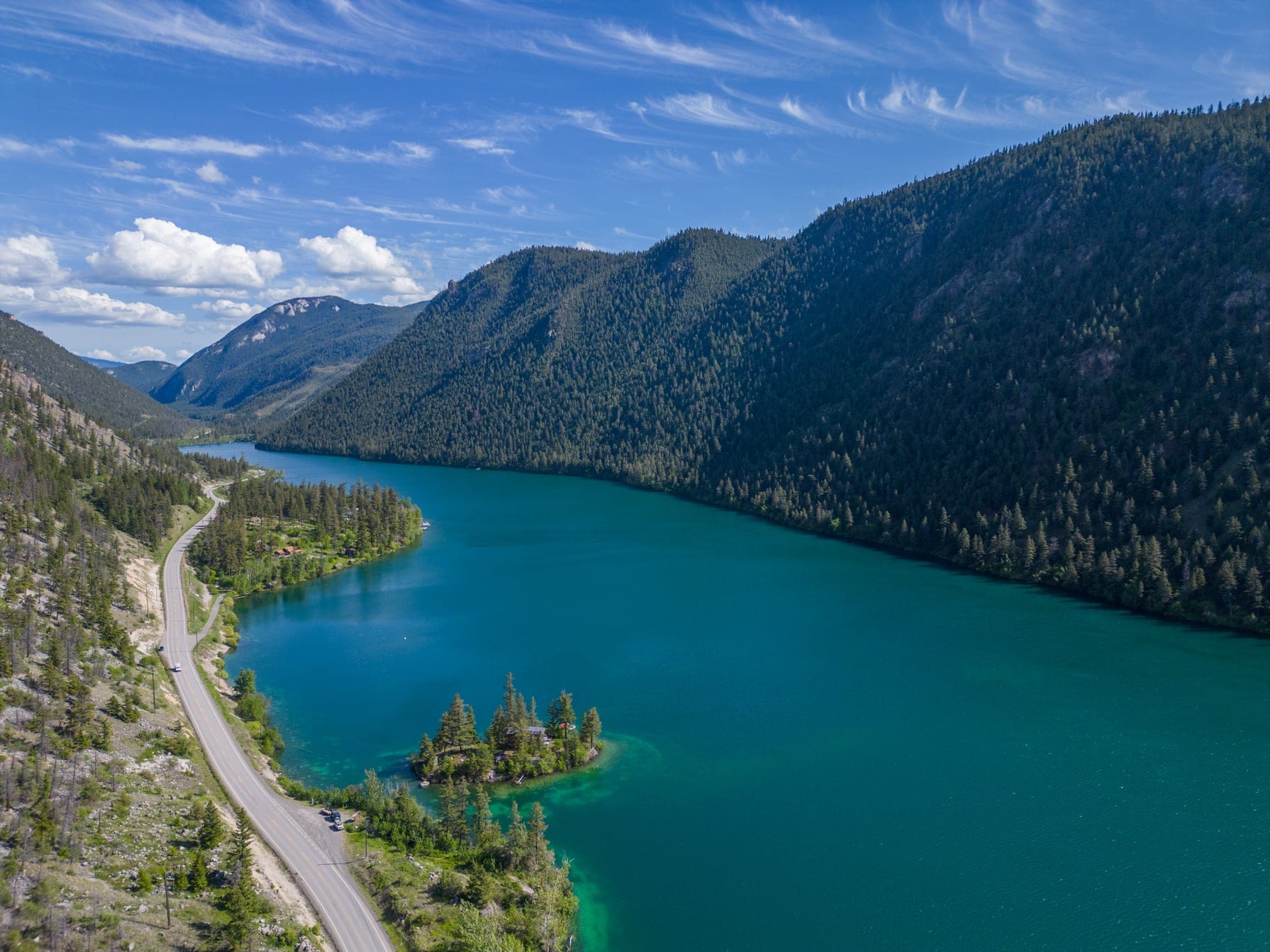 A small island sits in a large body of water, beside a highway on the left, facing a stunning view of forest covered mountains on the right.