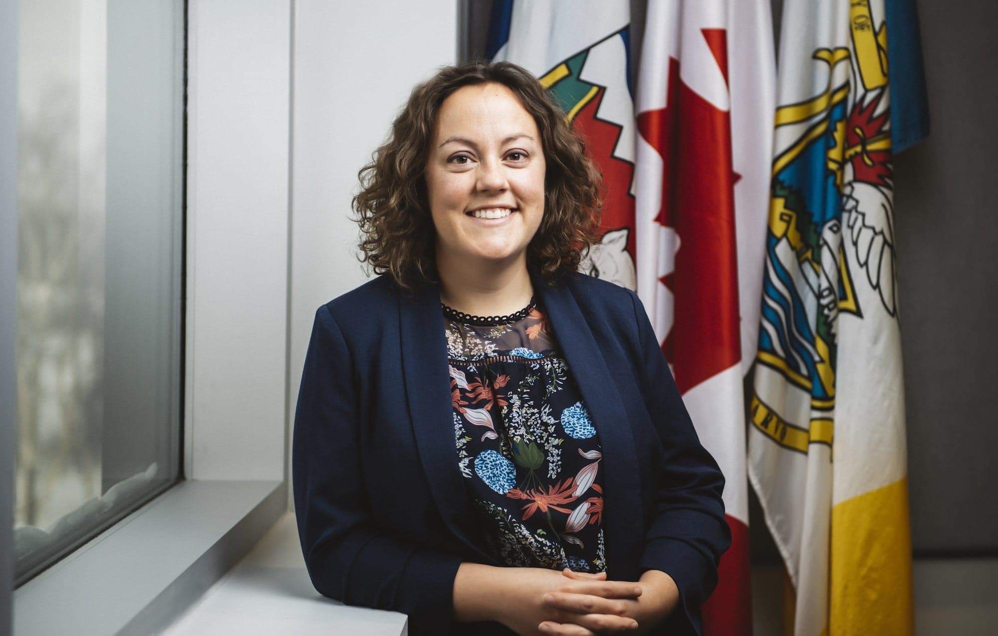 A woman in a blue blazer and colourful shirt stands smiling in front of the flags of the Northwest Territories and Canada.