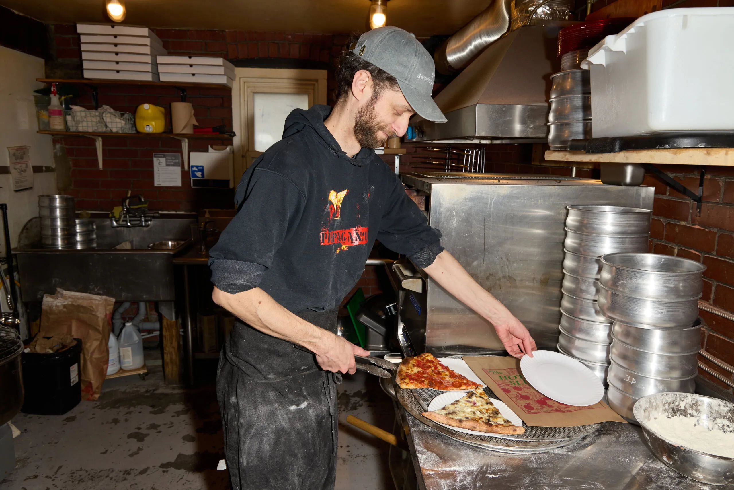 A man in a black hoodie and baseball cap putting a piece of pizza on a plate