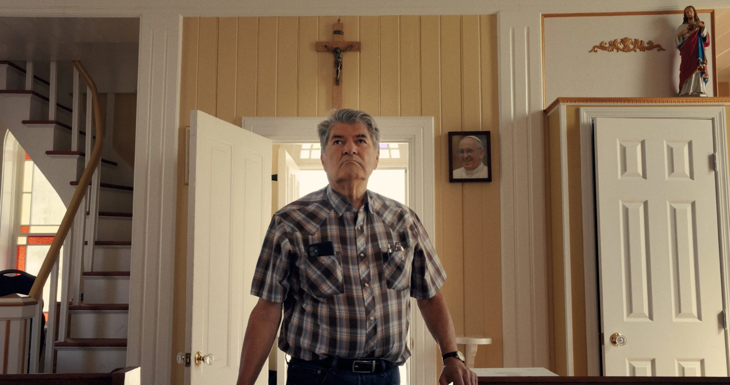 A man in a plaid shirt standing in an open doorway beneath a cross