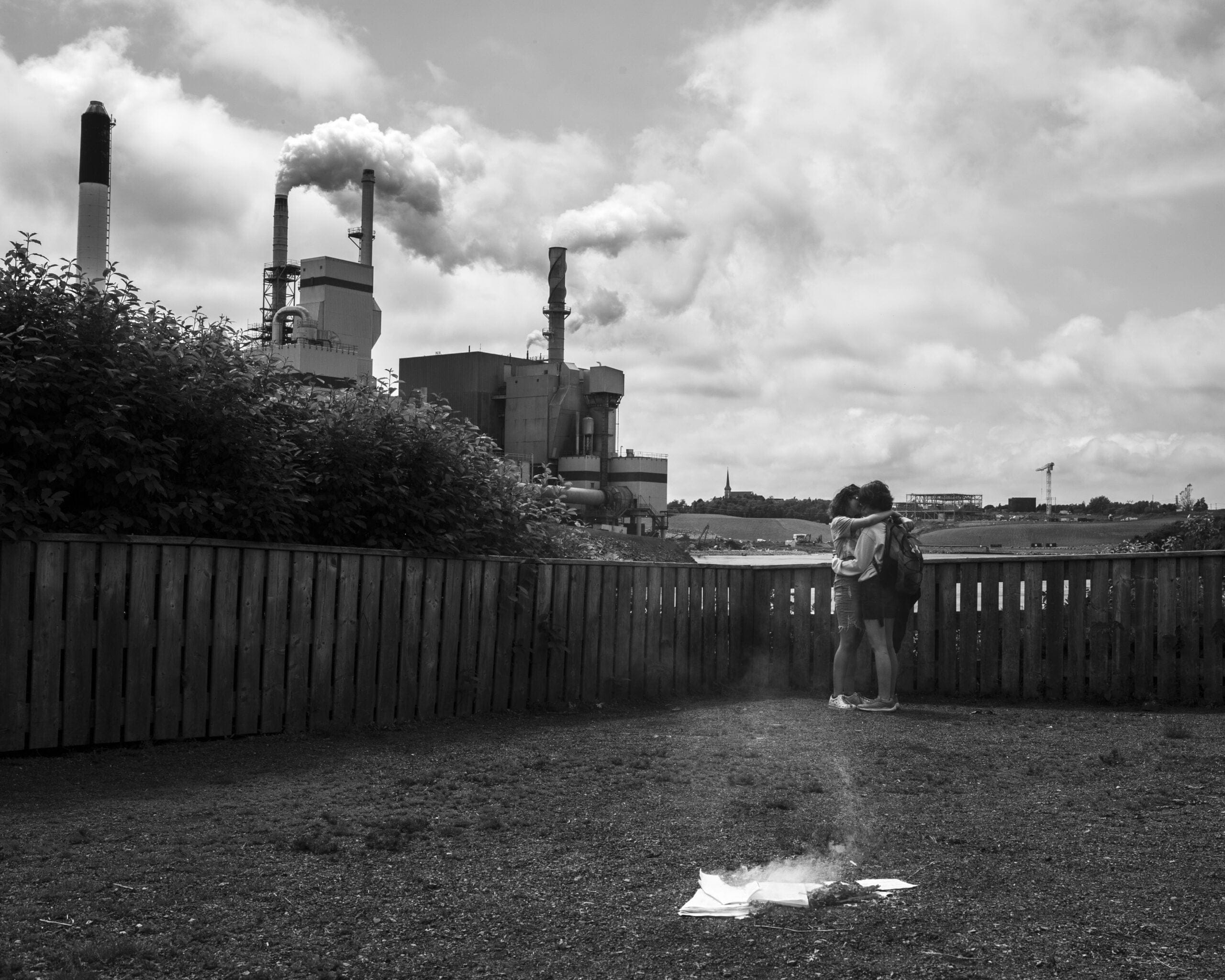 Two teenage girls hug in a backyard backing onto a large factory with smoke flowing out of smokestacks. There are papers burning on the ground on the lawn