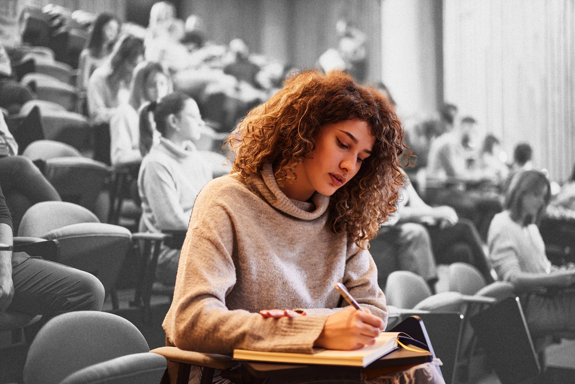A student in a beige turtleneck, sitting in a crowded classroom and writing in a notebook by hand