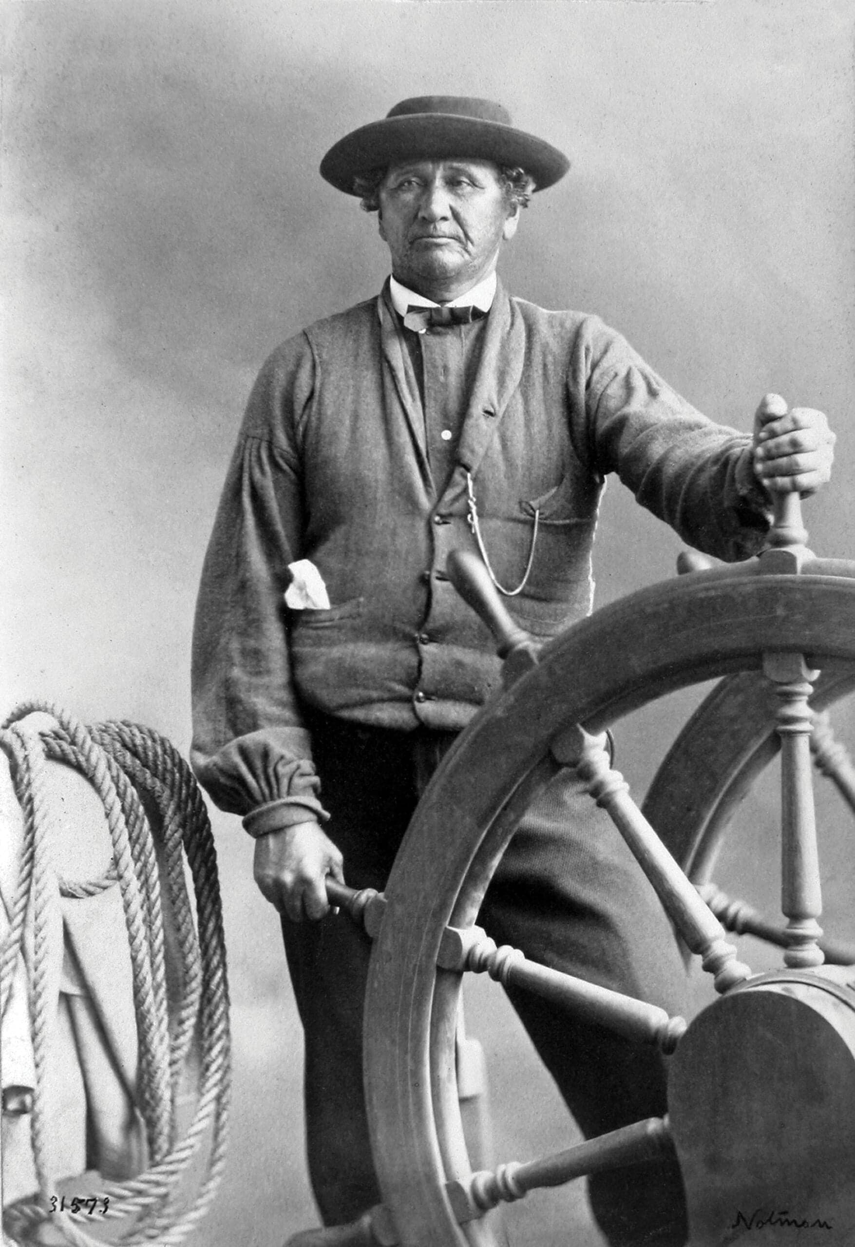 A man in a hat, holding a steering wheel in his left hand with a rope at his side