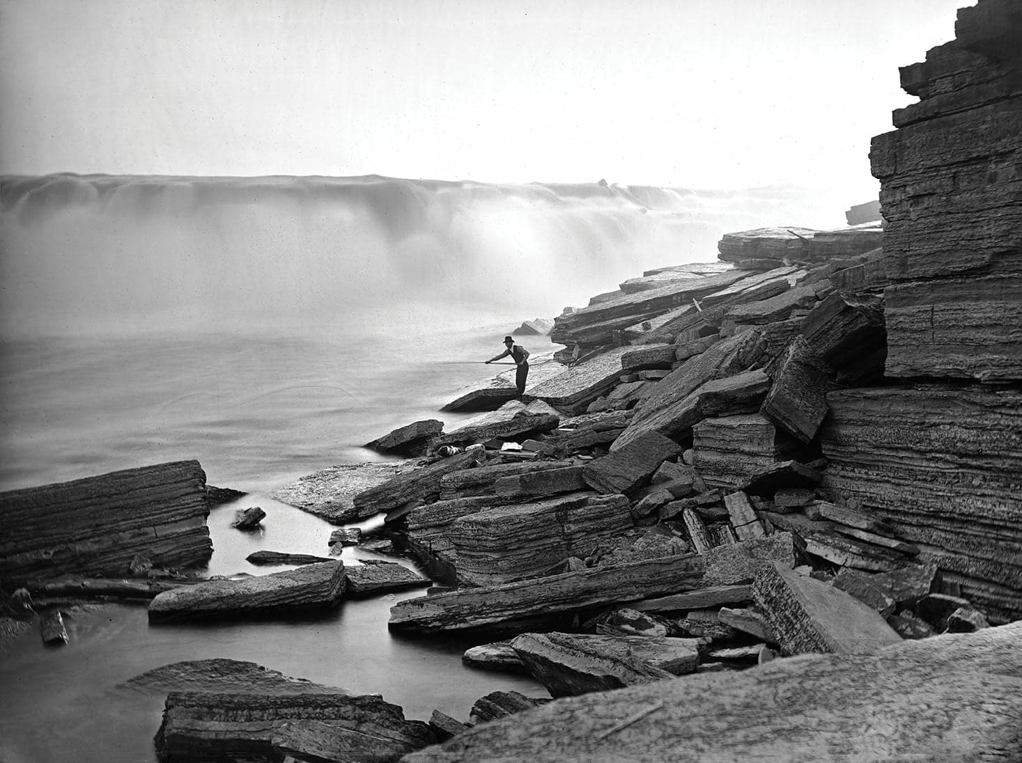 A man in a bowler hat holding a pole over the base of a waterfall