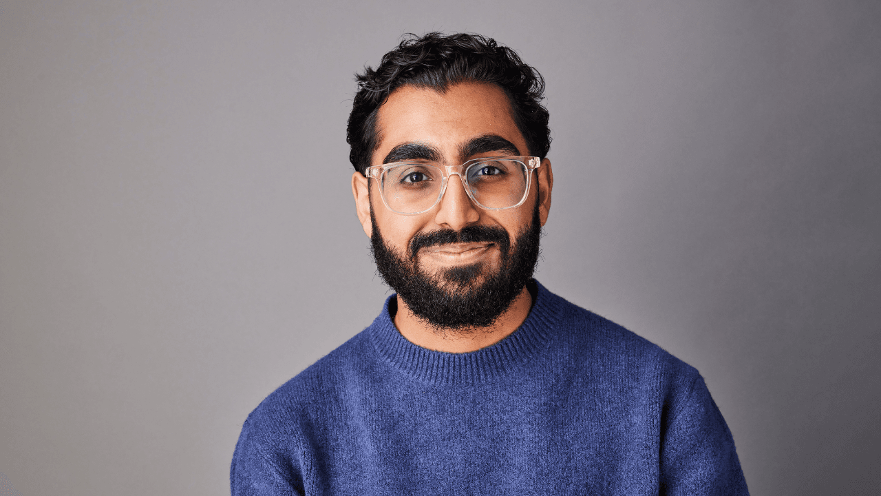 South asian man with a beard wearing a dark blue sweater and clear glasses in front of a dark grey backdrop.