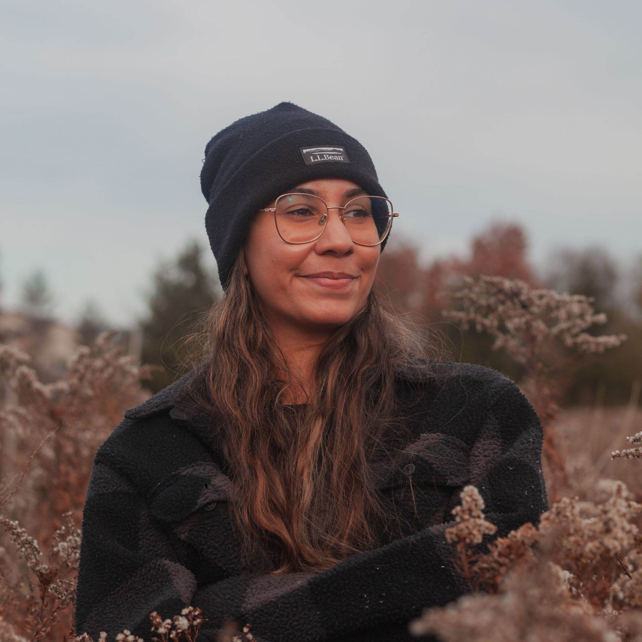 A woman in a black L.L. bean beanie, wire-rimmed glasses, and a black-and-brown checkered coat, standing in a field
