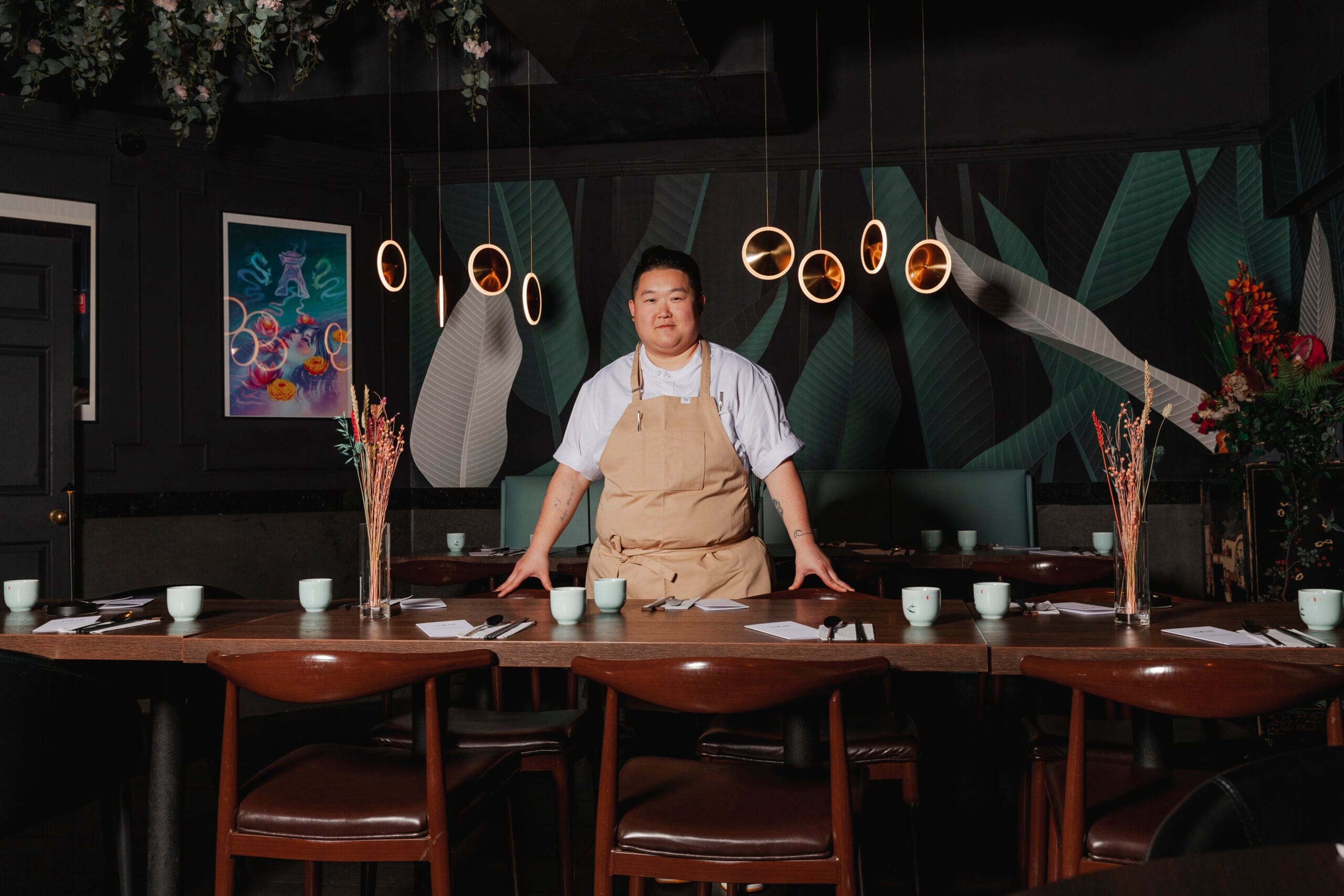 Chef Eva Chin wearing a white shirt under a tan apron, standing in a dark green dining room with a large wooden table in it