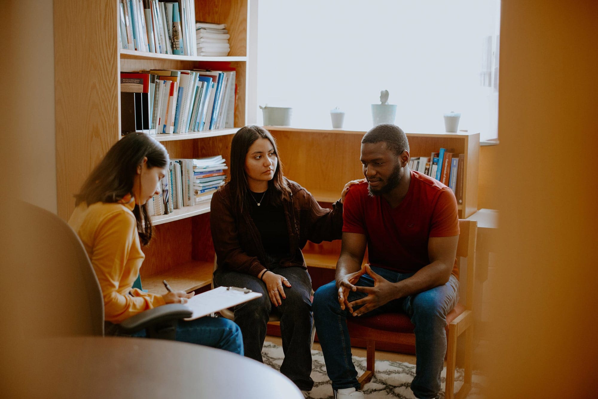 Three students sitting in a library, with a girl holding a man's shoulder as he looks down. The other student is writing on a clipboard across from them.