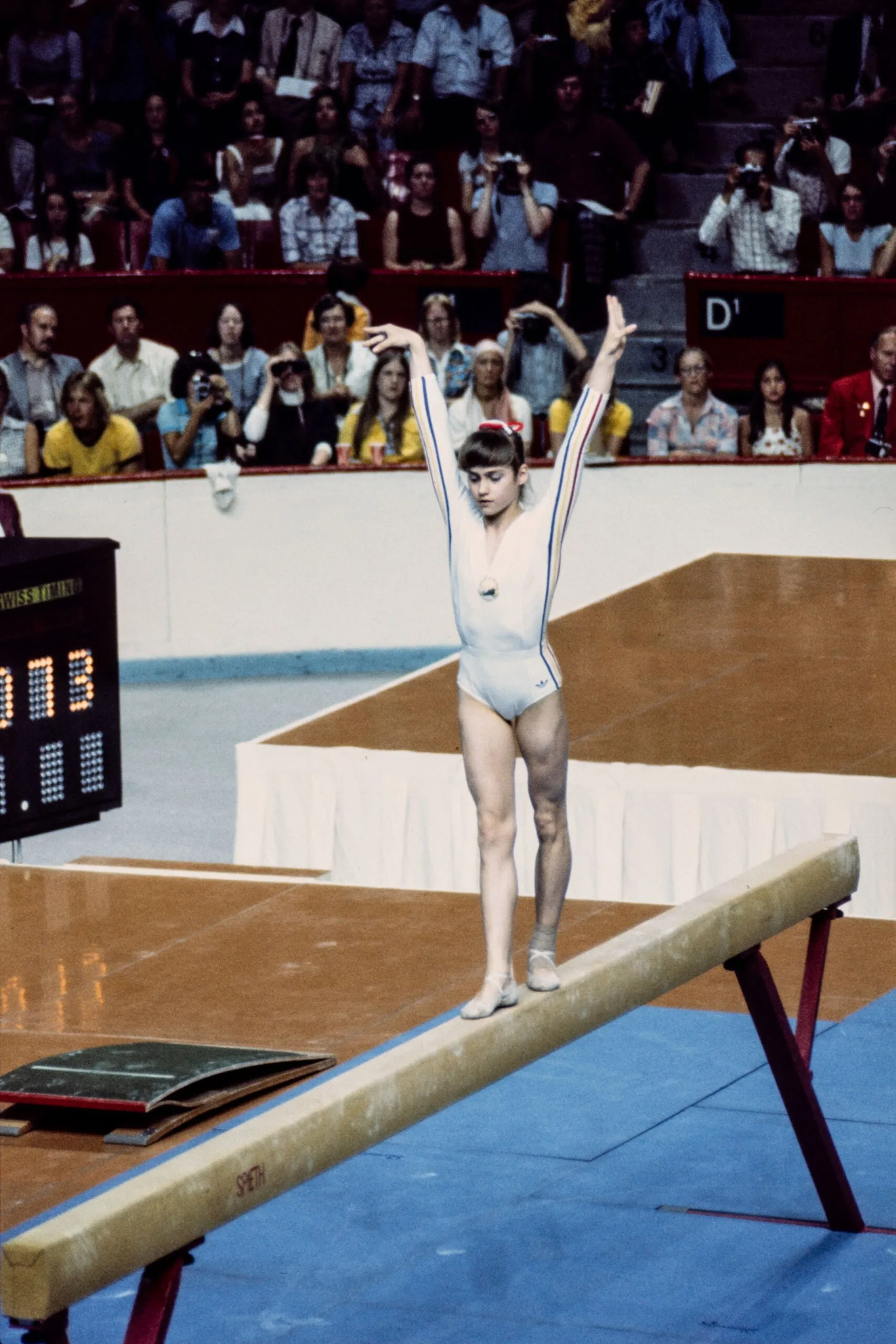 Nadia Comaneci in a white unitard with her arms raised in a V over her head, as she stands on the balance beam with a crowd behind her