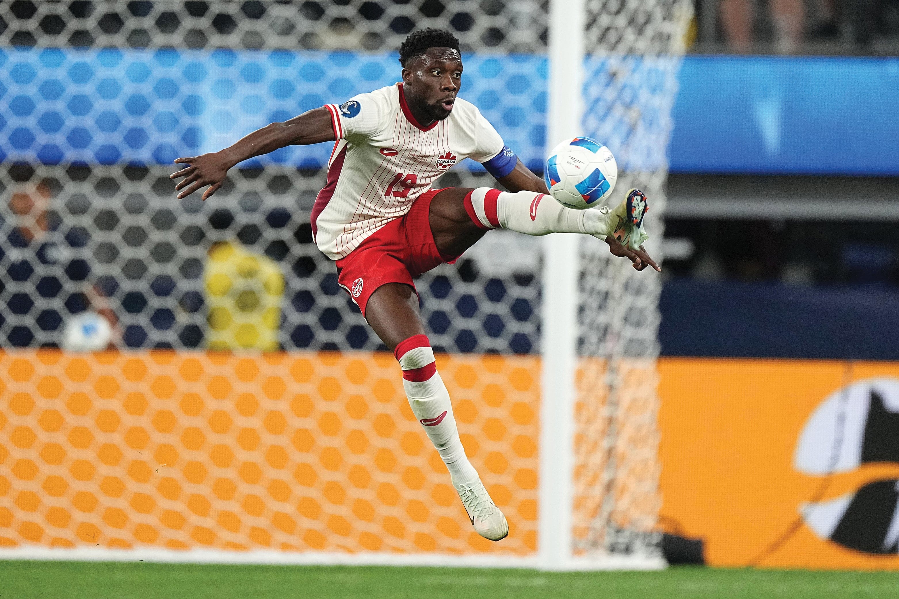 A soccer player (Alphonso Davies) is jumping in the air, catching a soccer ball with his left leg during a game. A white goal net behind him.