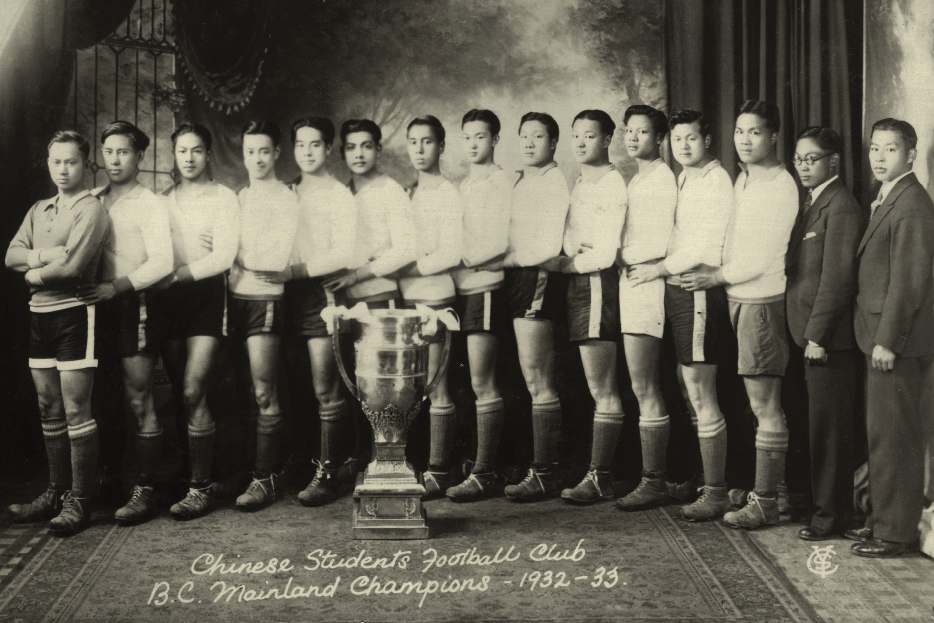 Sepia toned, black-and-white picture in which 15 men are lining up, posing for the picture. A huge, shiny trophy is put in front of them, and under that, a hand-written note says "Chinese students football club B.C. mainland champions - 1982-33."