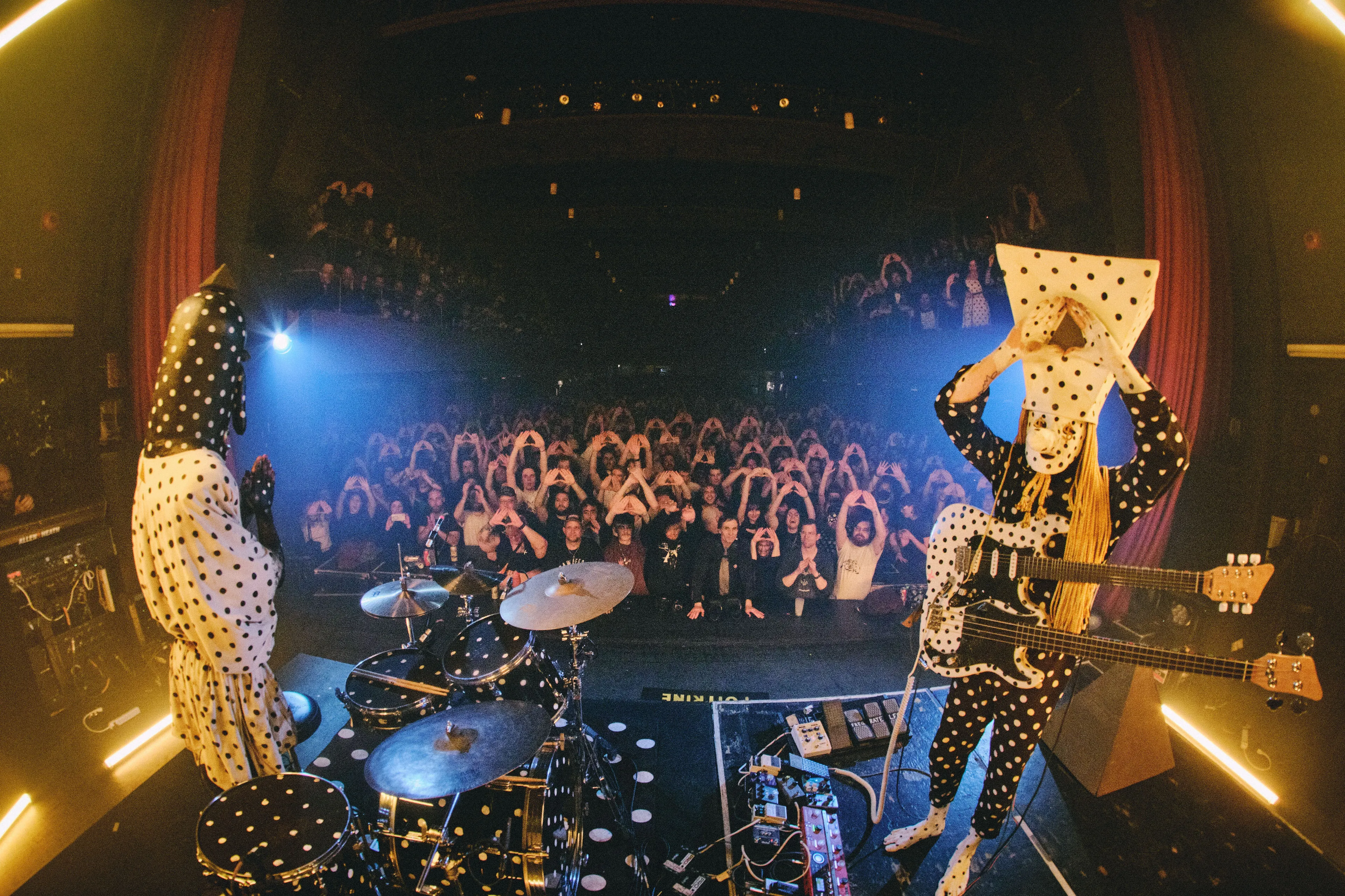 Two people (Angine de Poitrine) are performing on the stage with their costumes and instruments covered by black-and-white polka dots. The audiences are making triangle sign with both hands on their head. photographed with the fish eye lenz.