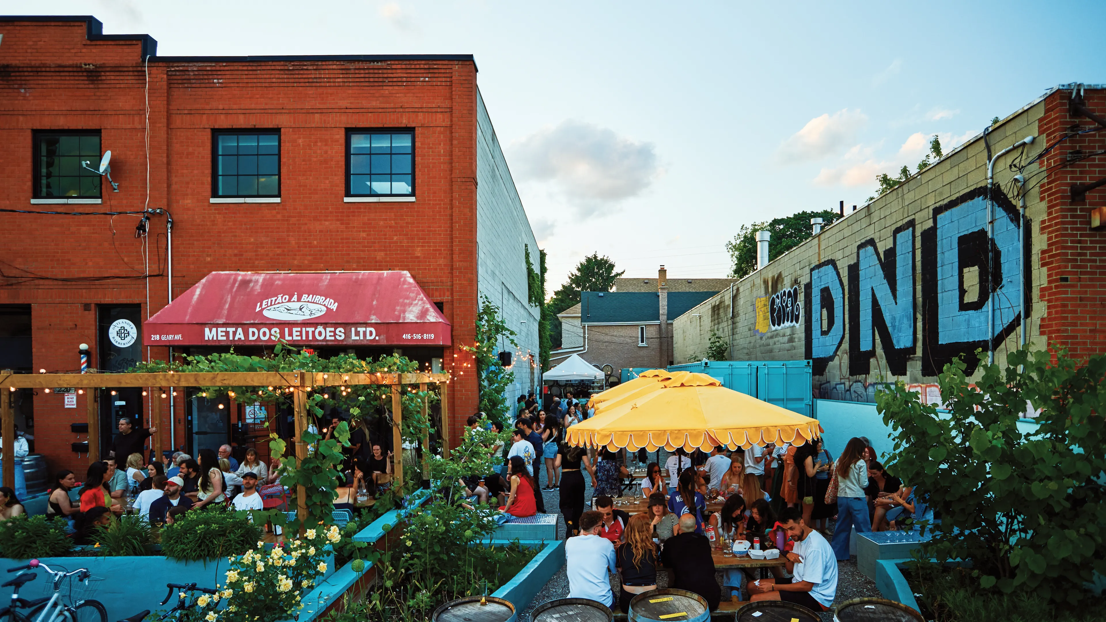 Restaurant's patio under the blue sky, surrounded by the brick wall, flower gardens and the barrels. People eating at tabes under the bright yellow umbrellas