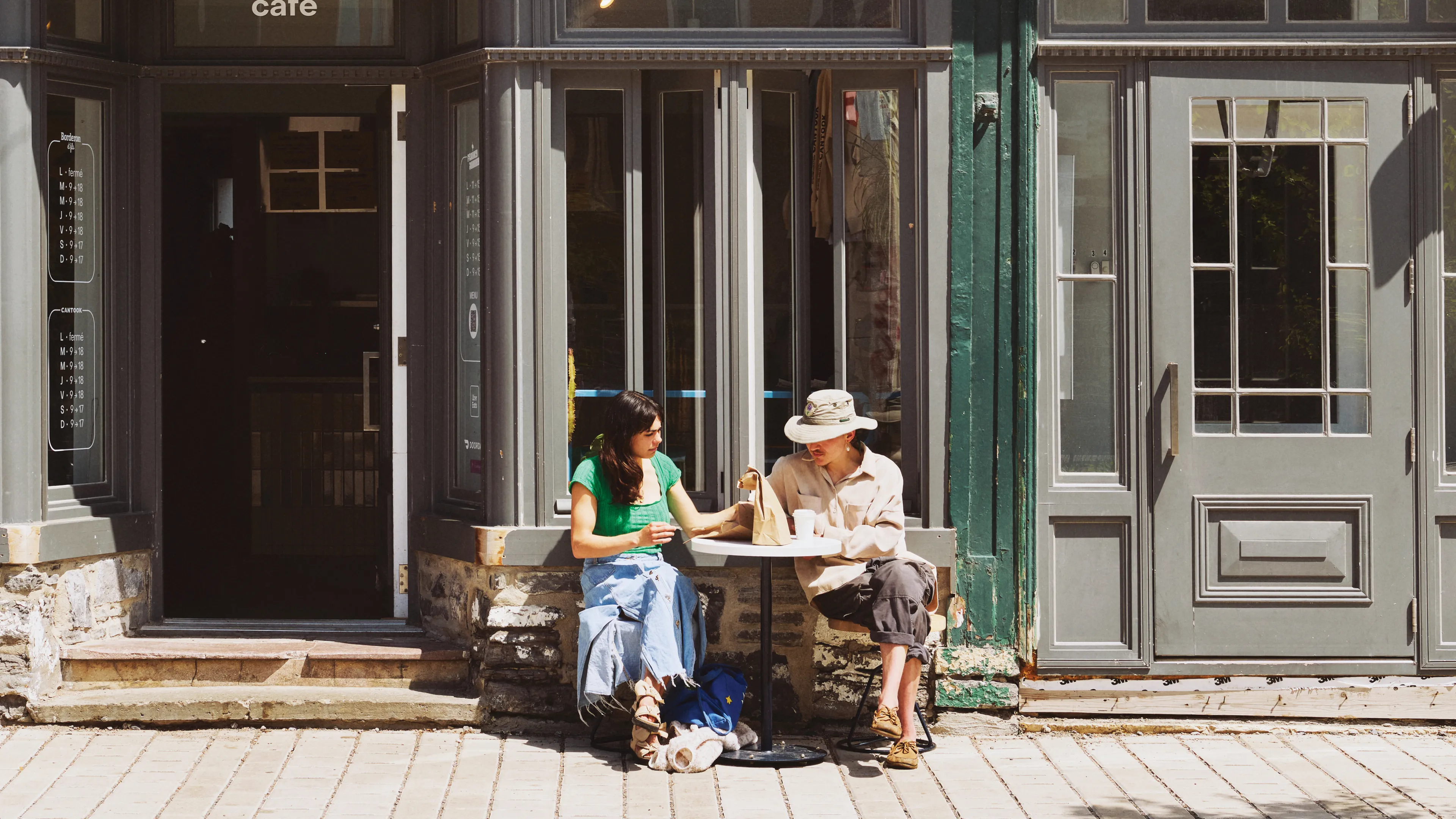 Two people sitting at patio of a cafe that says "sandwicherie" on its window