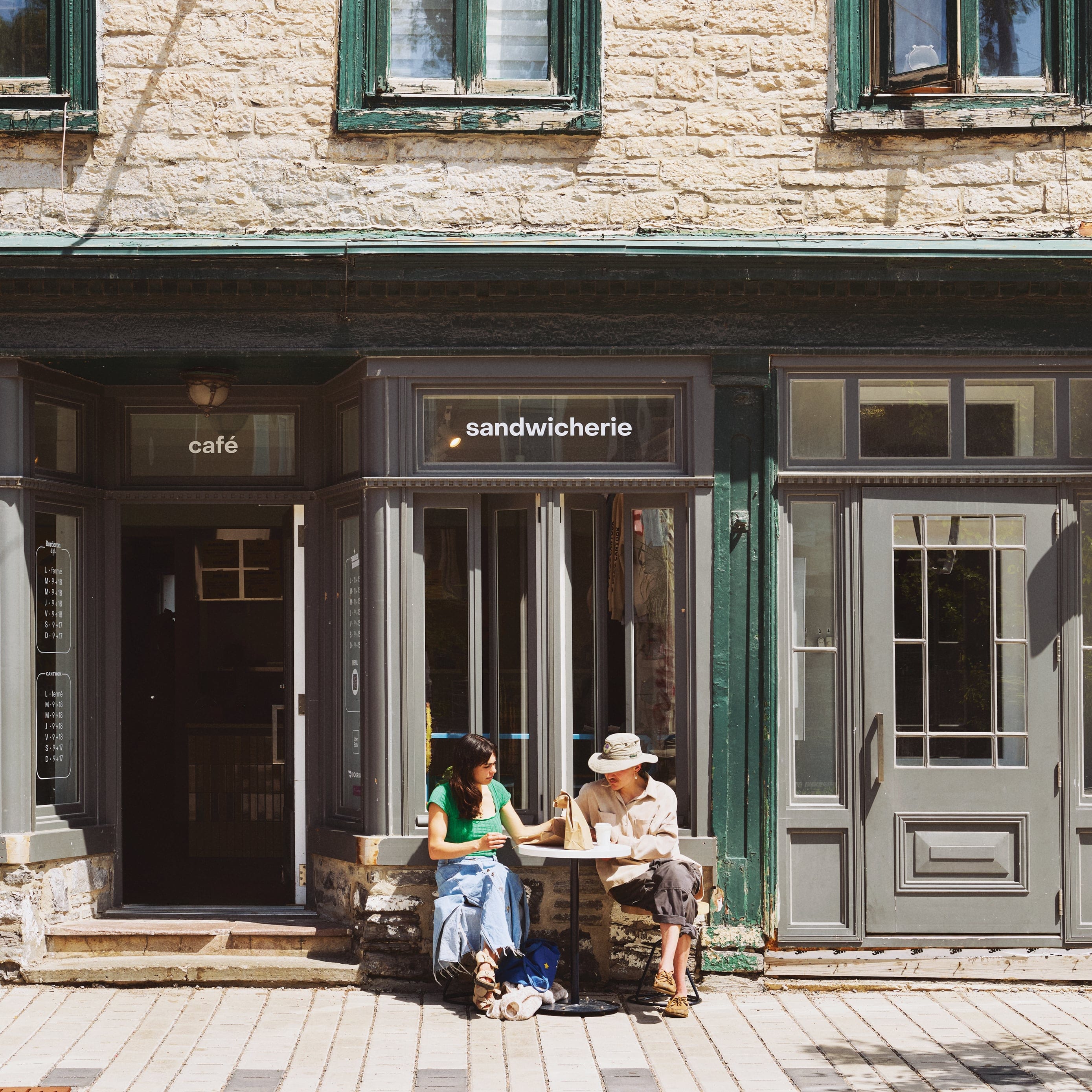 Two people sitting at patio of a cafe that says "sandwicherie" on its window