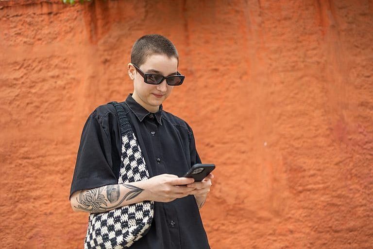 A teenager with short black hair, sunglasses, a buttoned up t-shirt and checkered purse staring at their phone. Pictured against an orange wall.