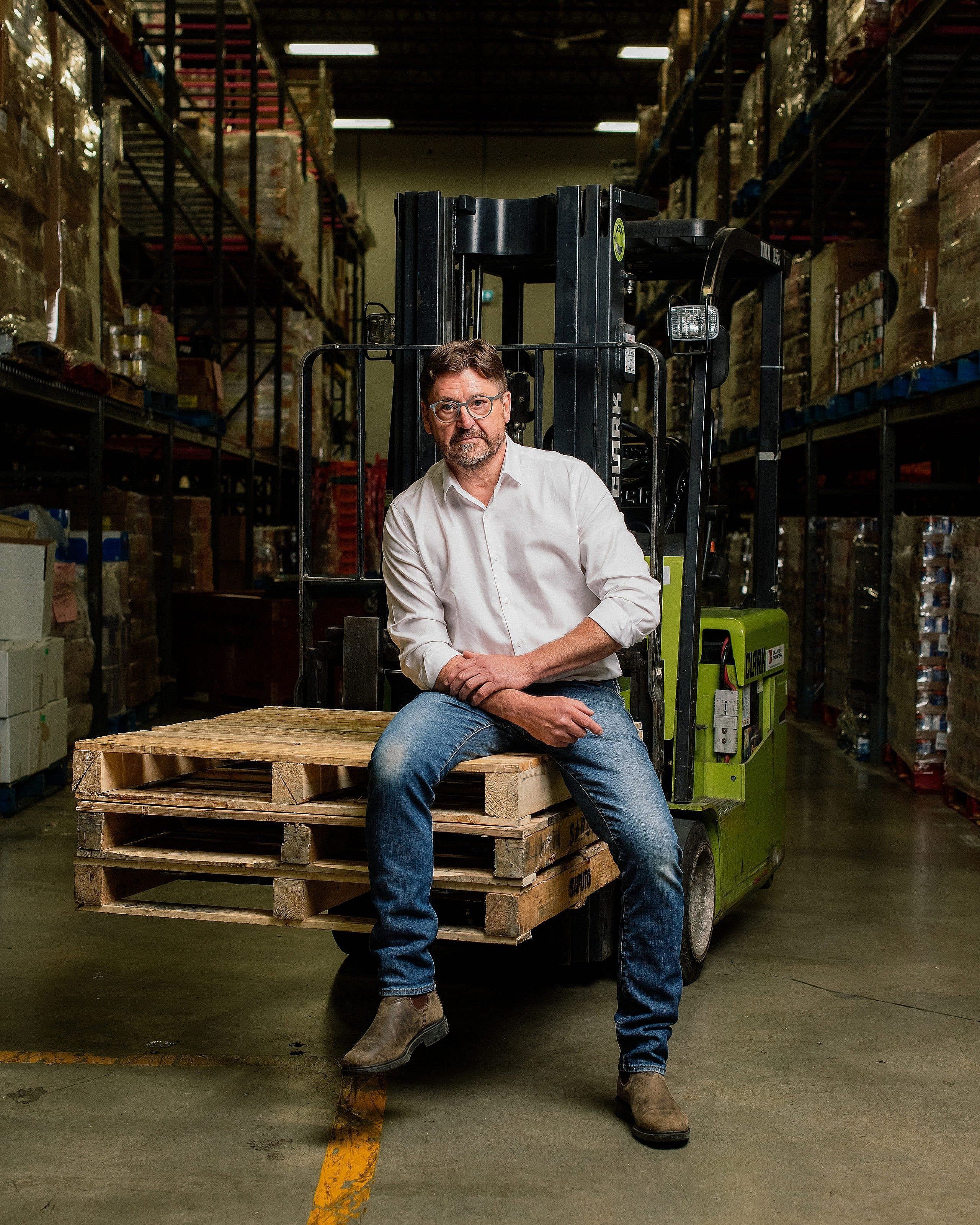 A man in a white button down shirt sits in a warehouse looking at the camera.