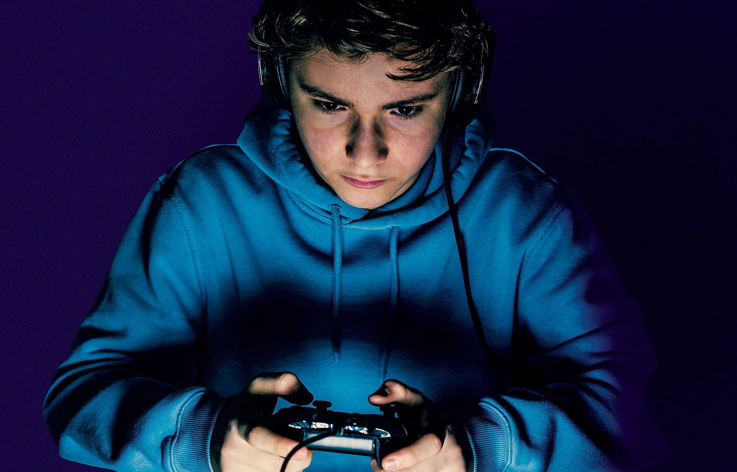 A boy in a royal blue hoodie stares intently at a screen ahead of him, holding a gaming console in his hands