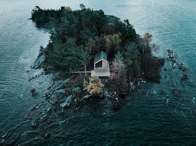A small cabin with a black roof sits on a isolated and forrested island surrounded by water.