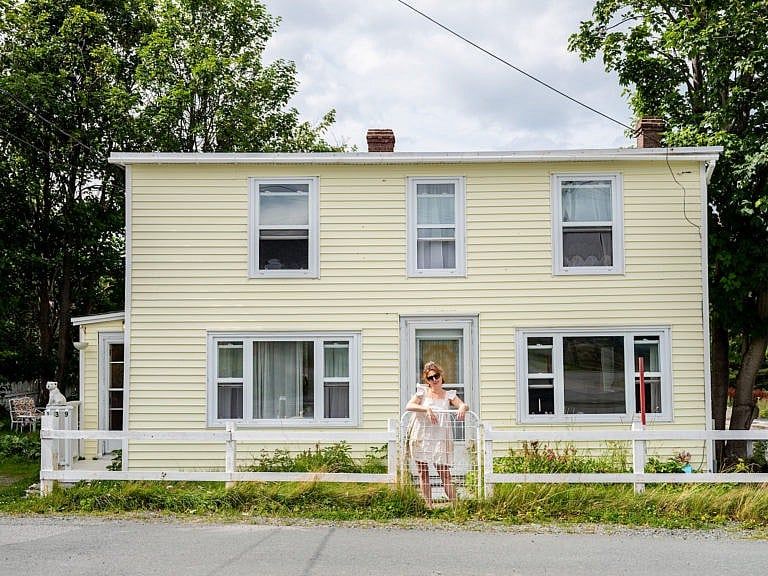 A woman leans against the gate of a white fence. She stands in front of a pale yellow house.