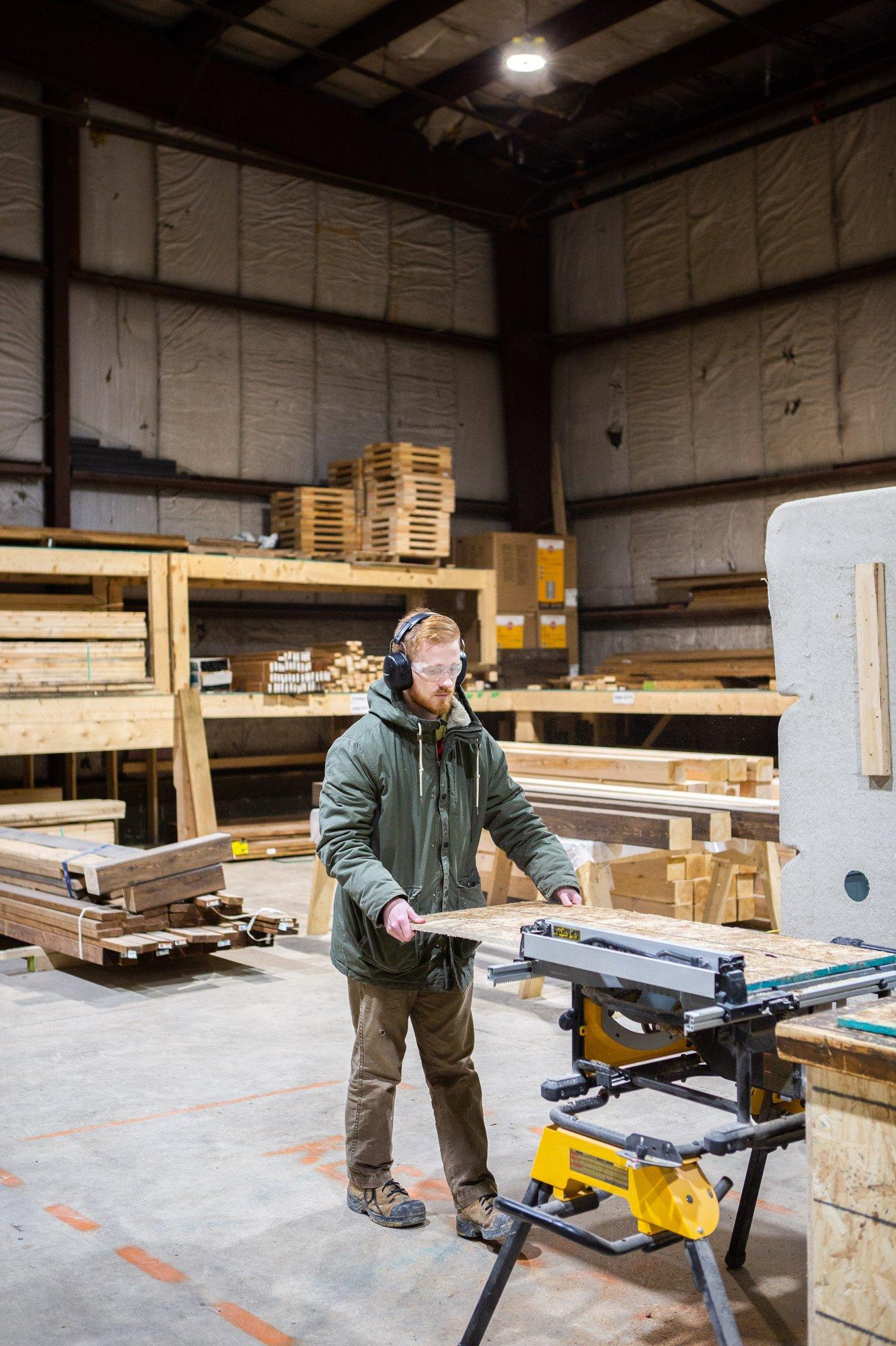 A photo of a man in a hoodie, wearing ear and eye protection and feeding wood through a machine