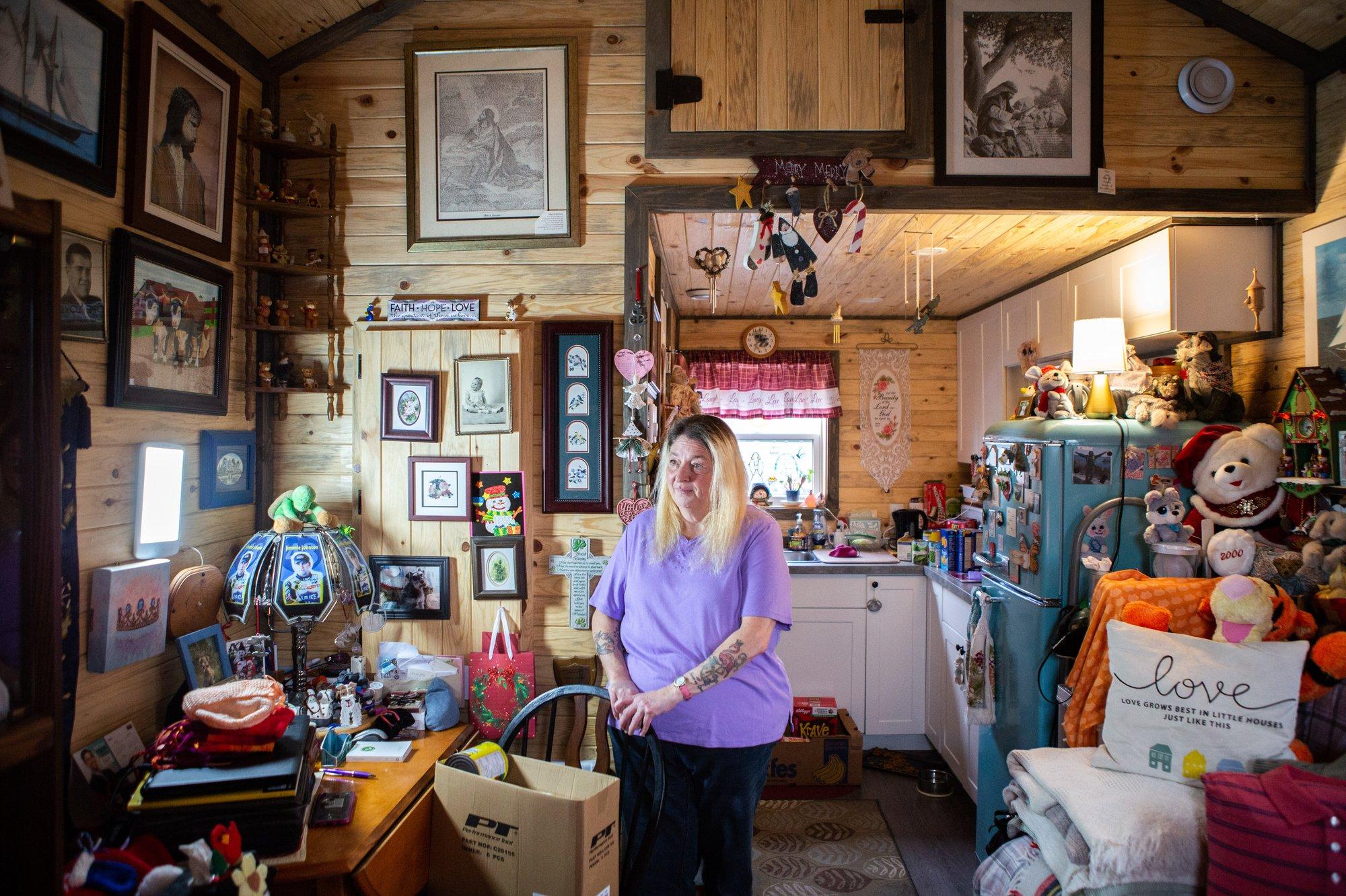 A woman in a purple sweater standing in a tiny home, with wooden walls covered in framed art